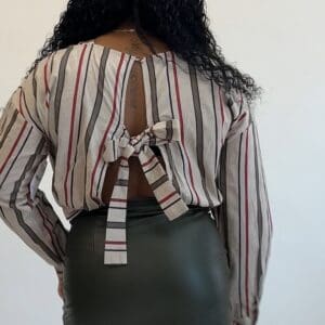 A person with long curly hair is wearing a striped shirt with an open back and bow detail, paired with a dark skirt. The background is plain white.