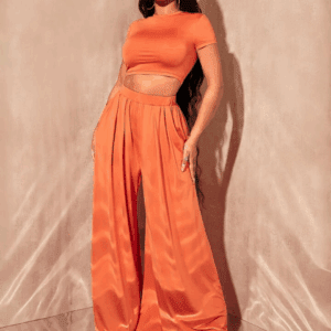 A woman models the 2PC Short Sleeve Crop Top w/ Wide Leg Pants set in orange, featuring a matching crop top and high-waisted wide-leg satin pants, posing with one hand in her pocket against a neutral background.