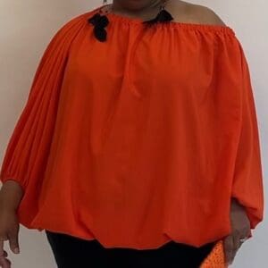 Person wearing the Puffy Shirt Dress—an off-the-shoulder, loose-fitting orange dress with long sleeves—paired with dark pants, standing against a plain light background.
