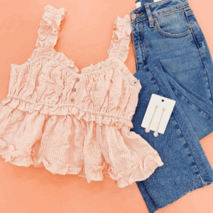 A Gingham Print Crop Top in pink, blue jeans, and gold earrings are arranged on a peach background.