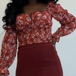 A person wearing a long-sleeved, red floral blouse with a square neckline and a high-waisted, textured red skirt, standing against a plain white background.