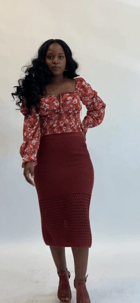A woman stands against a plain background wearing a red floral long-sleeve top, a red crochet midi skirt, and heeled sandals.
