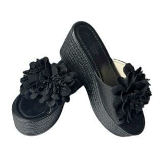 A pair of black platform sandals with large black fabric flowers on the straps, displayed against a white background.