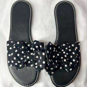 A pair of black slide sandals with white stitching and large black bows featuring white polka dots, placed on a white surface.