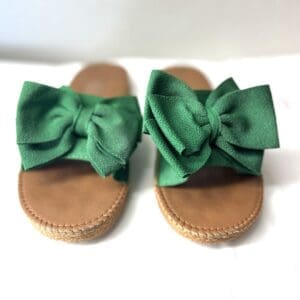 A pair of tan slide sandals with large green fabric bows on the straps displayed on a white surface.