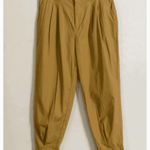 Sheer Cover Up Pants in tan, high-waisted with pleats and button-zip closure, displayed on a black hanger against a white textured wall.