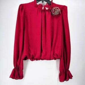 The Red Ruffle Wrist & Neck Blouse with Blign Rose features long sleeves, ruffled cuffs and neckline, and a decorative beaded rose brooch on the left shoulder, shown on a hanger against a white background.