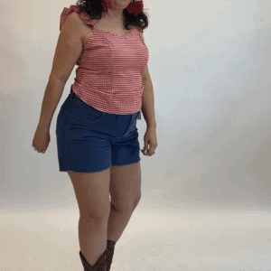 A person stands against a white background wearing the Gingham Top in red, paired with blue shorts, brown cowboy boots, and red earrings.