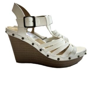 The Strappy Sandal is a white wedge with a black buckle, wooden platform heel, and metallic studs outlining the sole, shown from the side.