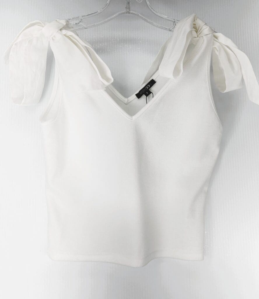 The White Tie Shoulder Top is a sleeveless V-neck with wide shoulder straps tied in bows, shown on a clear plastic hanger against a light background.
