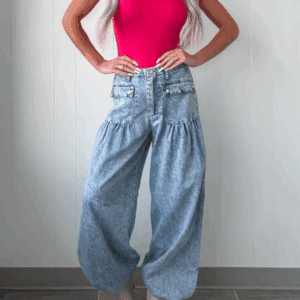 A person stands against a light-colored wall wearing a bright pink fitted short-sleeve top, 6 Button Pocket Dress Pants in a high-waisted baggy light-wash style, and beige platform boots.