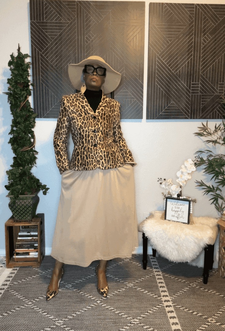 A person stands indoors wearing a leopard print blazer, beige skirt, beige hat, sunglasses, and a Wrap Belt. The background features plants, books, and geometric wall art.