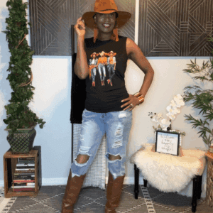 A woman stands indoors, wearing a brown hat, black graphic tank top, Denim Holey Jogger Overalls, and brown knee-high boots. She poses in a stylishly decorated room with plants and wall art.