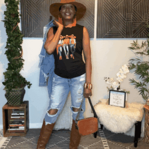 Indoors, a woman wears a wide-brim hat, graphic tank top, Denim Holey Jogger Overalls, brown knee-high boots, and holds a brown purse with plants and decor in the background.