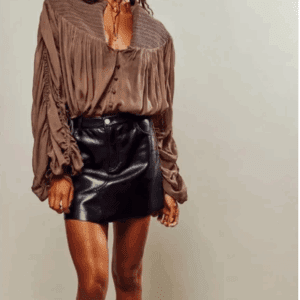 A person wearing a loose, brown, long-sleeve blouse paired with a black leather mini skirt stands against a plain background.