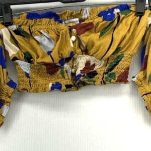 A Mustard Colored Crop Top featuring off-shoulder style, colorful abstract floral patterns, ruched elastic bands, and front buttons, displayed on a hanger.