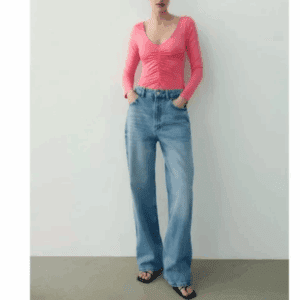 A person stands against a plain wall wearing the Off the Shoulder Be Bold Top in pink and light blue high-waisted wide-leg jeans with black sandals.