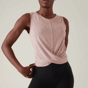 A person wearing a sleeveless light pink top with a front twist detail and black leggings, standing with one hand on their hip against a plain background.