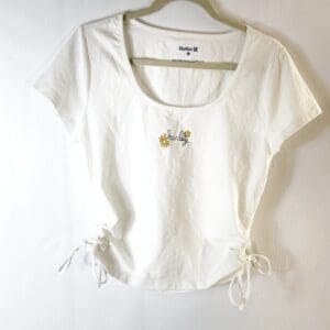The Hurley Side Drawstring Top is a white short-sleeve t-shirt featuring side drawstrings, daisy graphics, and a Hurley logo on the front, shown on a hanger against a white background.