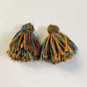 A pair of handmade tassels with multicolored yarn in shades of blue, green, brown, and gold on a white textured surface.