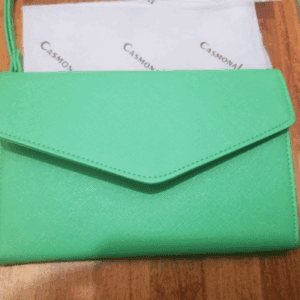A Green Wristlet rests on a wooden surface, with a branded Casmonal cloth partially visible behind it.