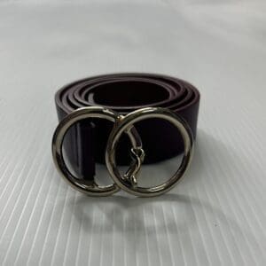 The Faux Leather Belt, coiled on a white textured surface, features a brown strap with two overlapping silver circular buckles.