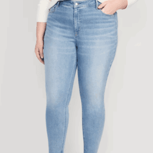 A person wearing a faux leather belt, light blue high-waisted skinny jeans, and a white sweater stands with one hand in a pocket.