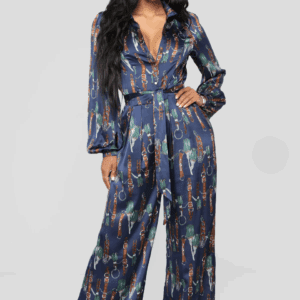 A woman models The Entertainer Satin Jumpsuit—a long-sleeve, deep V-neck, belted navy jumpsuit with a colorful chain and feather print—against a plain light gray background.