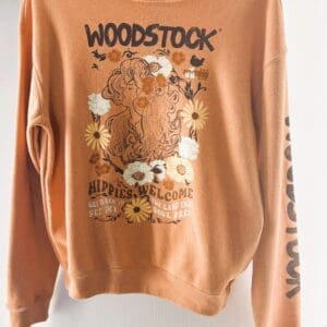The Woodstock Sweatshirt is peach-colored, features "WOODSTOCK" text, a floral design, and an illustration of a woman with daisies. It reads “HIPPIES WELCOME” and has additional text on the sleeve.
