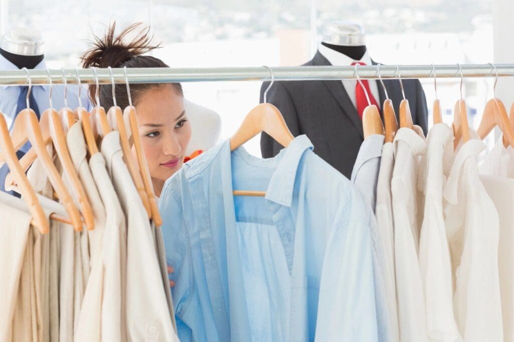A woman looks through a rack of light-colored shirts on hangers in a clothing store, with mannequins dressed in suits in the background.