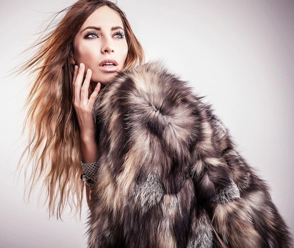 Woman with long, wavy hair wearing a large, multicolored fur coat, looking to the side with her hand resting on her face against a neutral background.