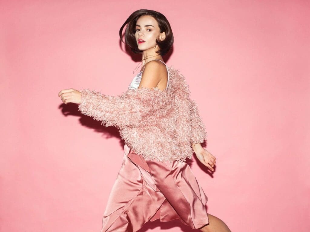 A woman in a pink fuzzy jacket and satin skirt walks in front of a solid pink background.