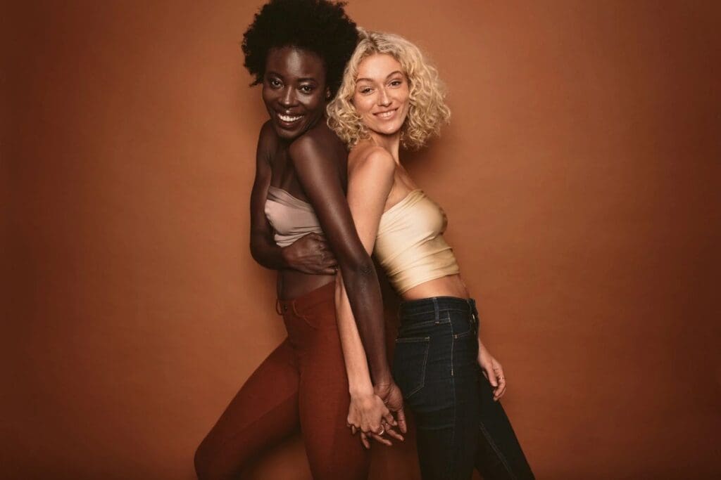 Two women stand side by side against a brown background, smiling and holding hands. One has dark skin and curly hair; the other has light skin and blonde curly hair.