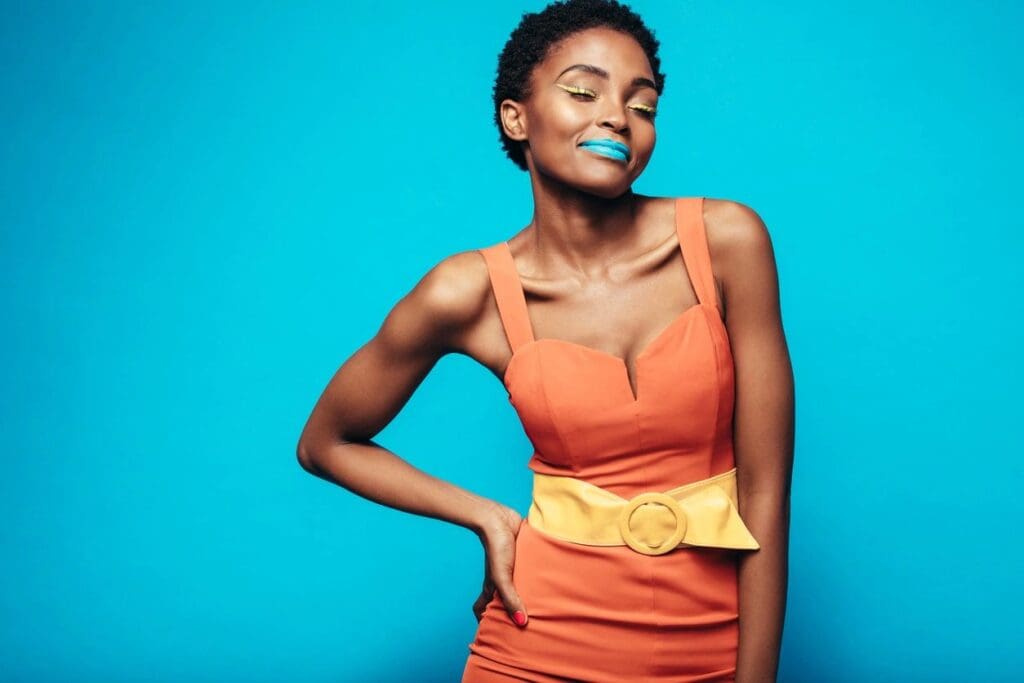 Woman in an orange dress with a wide yellow belt stands against a blue background, wearing blue lipstick and yellow eyeshadow, with one hand on her hip and eyes closed.