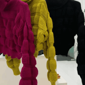 Three turtleneck crop knit sweaters in magenta, yellow-green, and black with raised, textured square patterns are hanging on a rack in a brightly lit room.
