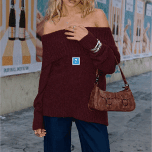 Person wearing an off-the-shoulder burgundy sweater, wide-leg jeans, silver bangles, and carrying a brown handbag, standing on a sidewalk with posters on a wall in the background.