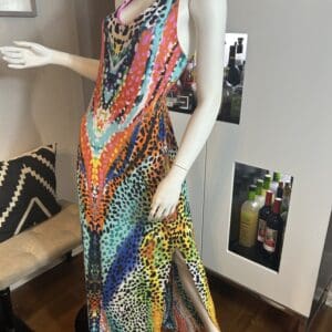Mannequin wearing a colorful, sleeveless, floor-length dress with a slit and a bold, multicolored dotted pattern, posed in a modern room near a bar cabinet.