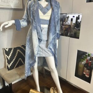 A mannequin dressed in a blue and white layered outfit with a striped skirt, standing next to a bar cabinet. Accessories include a wooden handbag and beige heels on the floor.