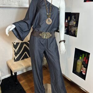 A mannequin dressed in a one-shoulder gray jumpsuit with a wide decorative belt, beaded necklace, and bangle, standing indoors near a bar cabinet.