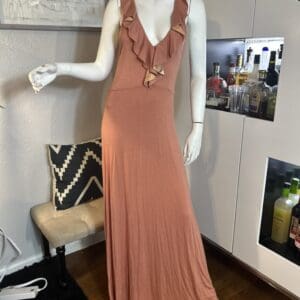 A mannequin displays a sleeveless, floor-length, rust-colored dress with ruffle accents on the neckline, set in a modern room with a bar cabinet and a small bench.