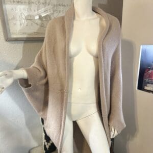 Mannequin wearing a loose, light beige open-front cardigan with wide sleeves, displayed indoors against a neutral wall with framed pictures in the background.