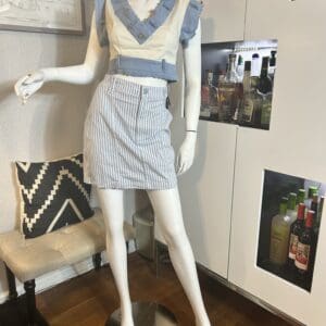 A mannequin dressed in a blue and white ruffled crop top and a blue and white striped skirt stands on a wooden floor next to a bar cabinet and a cushioned bench.