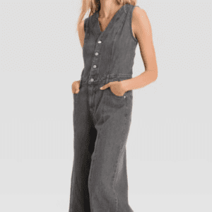 A woman wears Product, a sleeveless button-up dark gray denim jumpsuit with wide legs and side pockets, standing with her hands in the pockets and smiling.