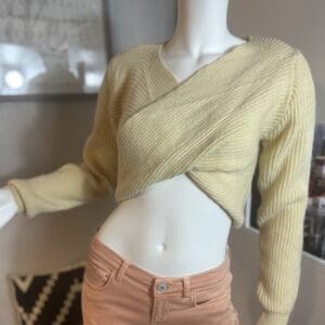 Mannequin dressed in a pale yellow ribbed wrap sweater and light pink pants, displayed in a room with framed art and a patterned pillow in the background.