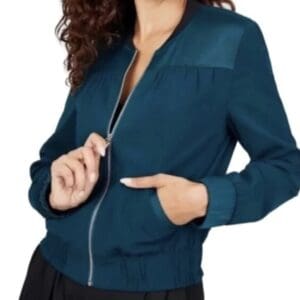 A woman wearing the Sabie Bomber Jacket, a dark teal zip-up style with long sleeves and gathered cuffs, stands with one hand in her pocket.