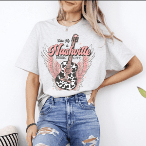 Woman wearing a light gray t-shirt with a graphic of a winged guitar and the text “Take Me to Nashville Music City,” paired with ripped jeans.
