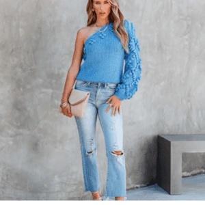 Woman stands against a gray textured wall, wearing the Rust One Shoulder Juniper Sweater (size S), ripped light-wash jeans, white heels, and holding a beige clutch.
