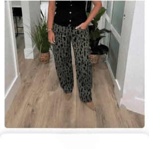 Person wearing patterned wide-leg pants, a black button-up top, and beige pointed shoes standing indoors on wood flooring near green potted plants.