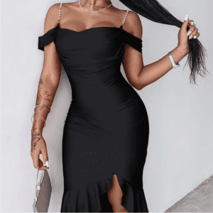A woman wearing an off-the-shoulder black dress with thin straps and a front slit stands against a white wall, holding a clutch bag.