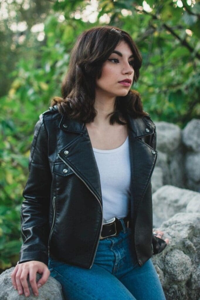 A woman with shoulder-length dark hair wearing a black leather jacket, white top, and blue jeans stands outdoors near rocks and greenery.